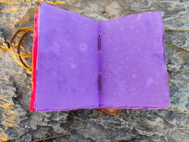 May include: A purple handmade journal with a red cover and brown leather binding. The journal is open to reveal blank pages.
