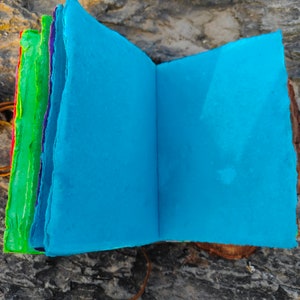 May include: A handmade journal with a blue cover and colorful pages. The journal is open to reveal the blank pages inside. The journal is sitting on a piece of wood.