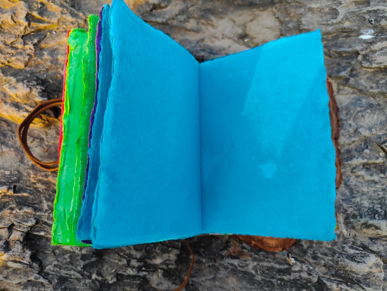 May include: A handmade journal with a blue cover and colorful pages. The journal is open to reveal the blank pages inside. The journal is sitting on a piece of wood.