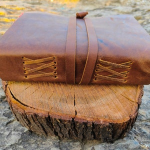 May include: A brown leather-bound journal with a leather strap closure and decorative stitching. The journal is resting on a wooden log.