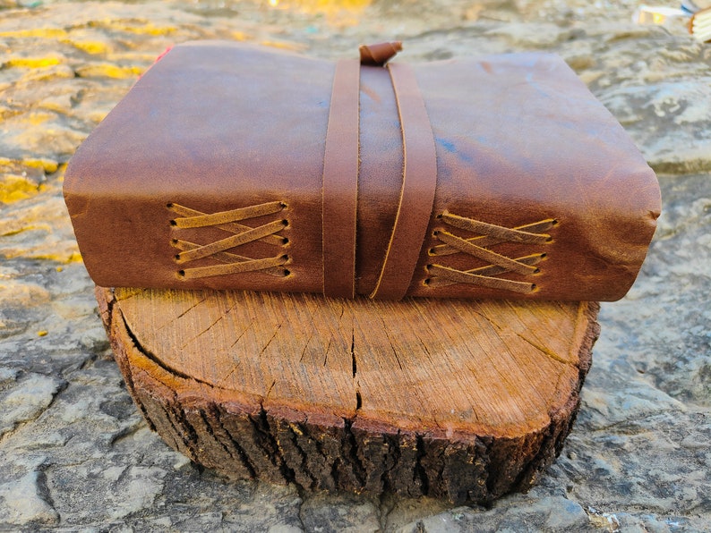 May include: A brown leather-bound journal with a leather strap closure and decorative stitching. The journal is resting on a wooden log.