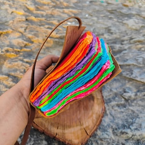 May include: A colorful handmade journal with a leather cover and a rainbow of paper pages. The journal is bound with a brown leather strap.