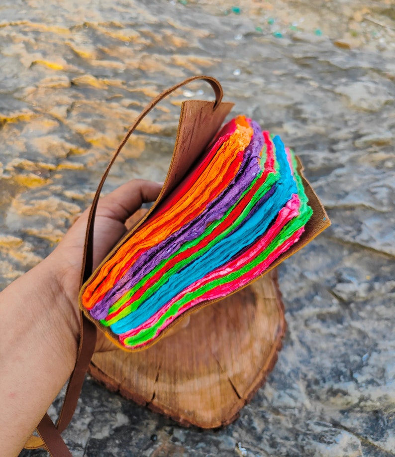 May include: A colorful handmade journal with a leather cover and a rainbow of paper pages. The journal is bound with a brown leather strap.