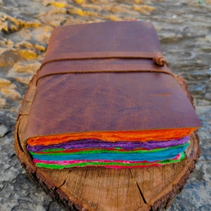 May include: A brown leather-bound journal with a colorful layered paper interior. The journal is closed and sitting on a wooden surface.