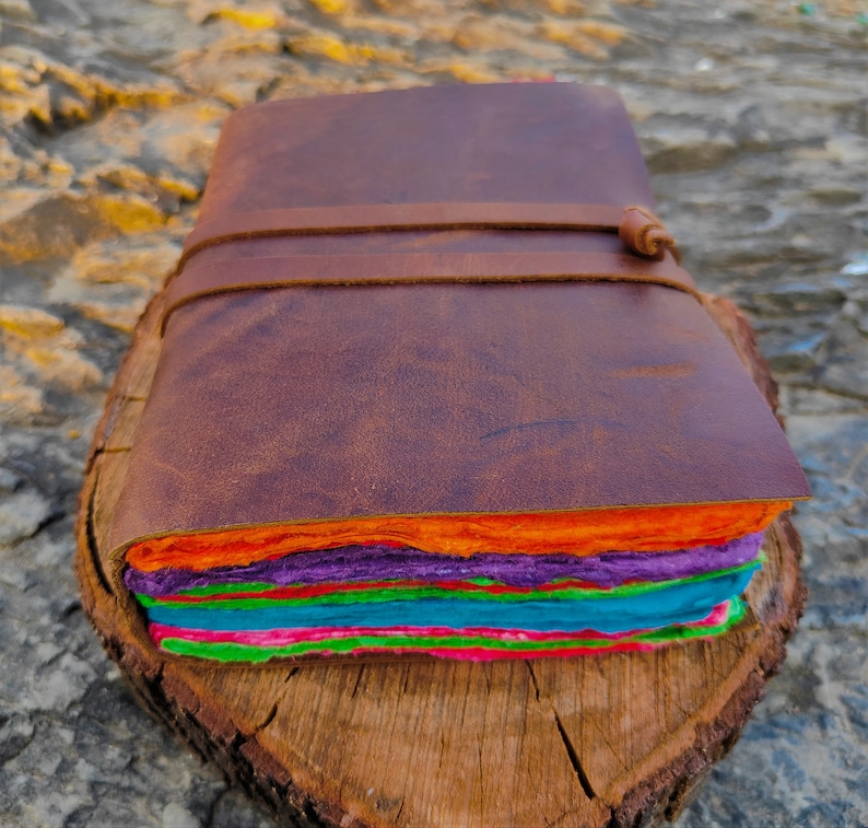 May include: A brown leather-bound journal with a colorful layered paper interior. The journal is closed and sitting on a wooden surface.