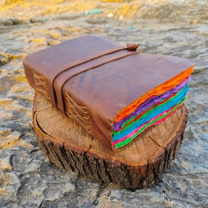 May include: A brown leather-bound journal with a colorful handmade paper interior. The journal is closed and has a leather strap with a buckle. It is sitting on a wooden stump.