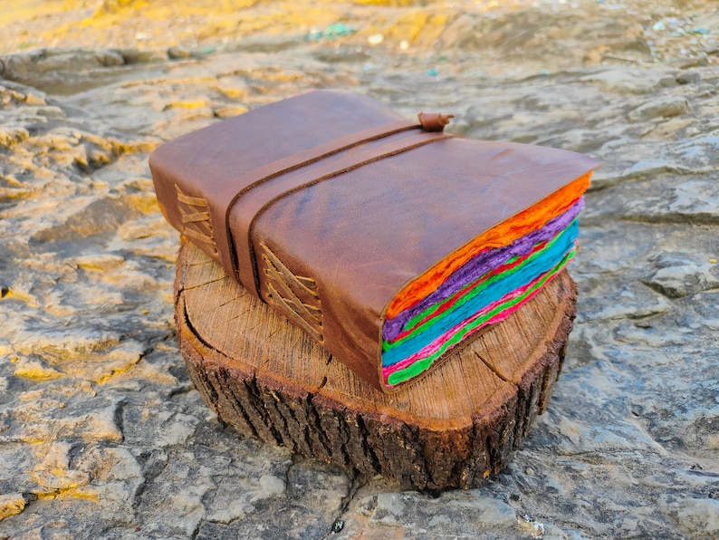 May include: A brown leather-bound journal with a colorful handmade paper interior. The journal is closed and has a leather strap with a buckle. It is sitting on a wooden stump.