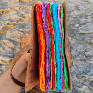 May include: A colorful handmade journal with a leather cover and a strap. The pages are made of recycled paper and are layered in shades of orange, red, green, blue, and purple.