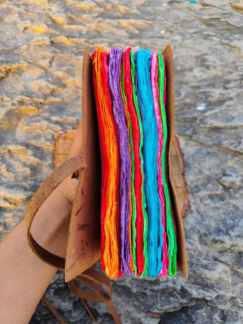 May include: A colorful handmade journal with a leather cover and a strap. The pages are made of recycled paper and are layered in shades of orange, red, green, blue, and purple.