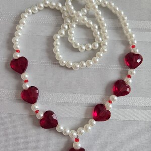 May include: A pearl necklace featuring red heart-shaped pendants. The necklace has a strand of white pearls with red heart-shaped accents and small red beads. The necklace is a romantic accessory.