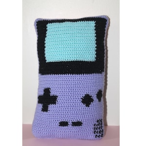 May include: A purple crocheted pillow shaped like a Game Boy with a teal screen and black buttons.