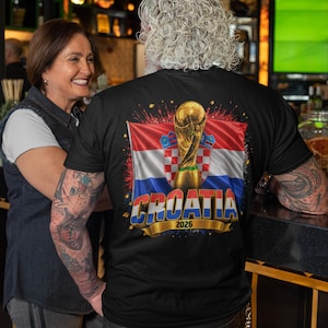 Black t-shirt worn by a male model featuring a Croatia football-themed design with the FIFA World Cup trophy, Croatian flag, and bold “CROATIA 2026” text on the front.