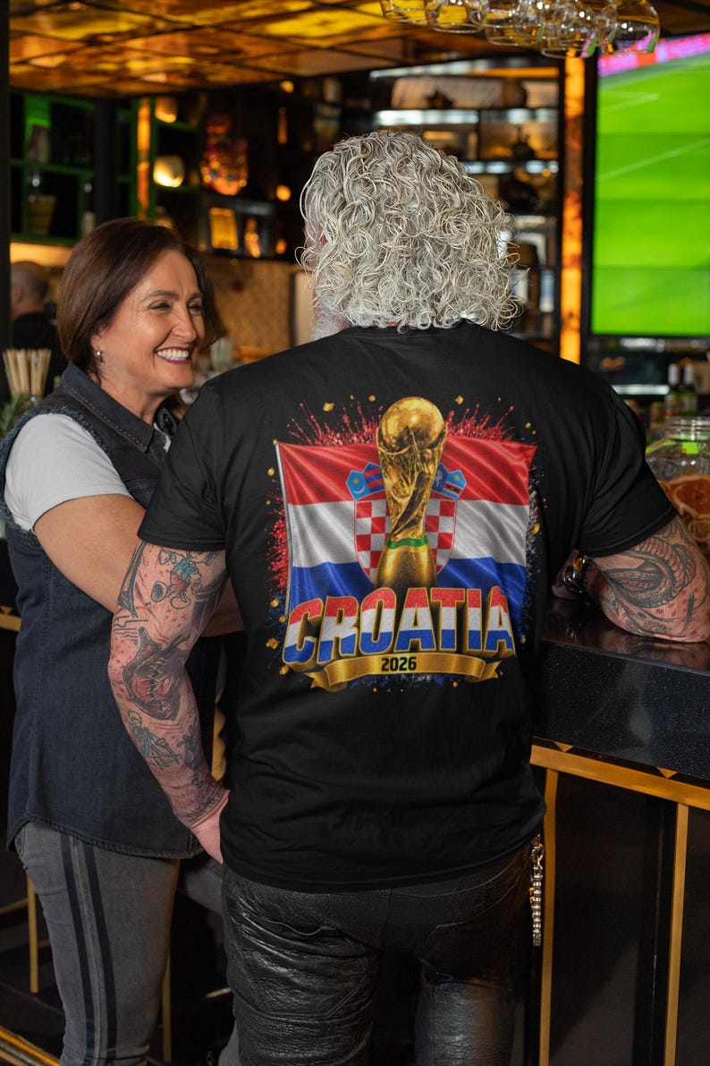 Black t-shirt worn by a male model featuring a Croatia football-themed design with the FIFA World Cup trophy, Croatian flag, and bold “CROATIA 2026” text on the front.