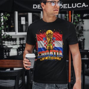 Black t-shirt worn by a male model featuring a Croatia football-themed design with the FIFA World Cup trophy, Croatian flag, and bold “CROATIA 2026” text on the front.