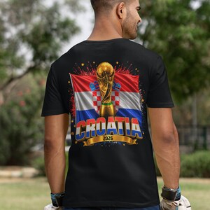 Black t-shirt worn by a male model featuring a Croatia football-themed design with the FIFA World Cup trophy, Croatian flag, and bold “CROATIA 2026” text on the front.