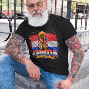 Black t-shirt worn by a male model featuring a Croatia football-themed design with the FIFA World Cup trophy, Croatian flag, and bold “CROATIA 2026” text on the front.