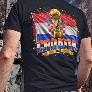 Black t-shirt worn by a male model featuring a Croatia football-themed design with the FIFA World Cup trophy, Croatian flag, and bold “CROATIA 2026” text on the front.