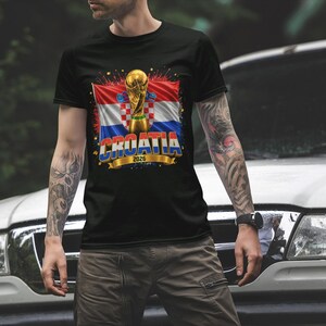 Black t-shirt worn by a male model featuring a Croatia football-themed design with the FIFA World Cup trophy, Croatian flag, and bold “CROATIA 2026” text on the front.