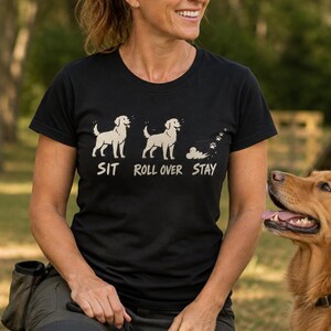 May include: A black short-sleeved t-shirt featuring a graphic of a light-colored dog in three positions with the text SIT, ROLL OVER, and STAY printed below. The first dog stands, the second stands, and the third shows paw prints and dust.
