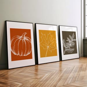 May include: Three framed illustrations of fall foliage. The first frame features a pumpkin in orange with a white outline on a rust-colored background. The second frame features a maple leaf in white on a yellow background. The third frame features a branch of berries in white on a dark brown background.