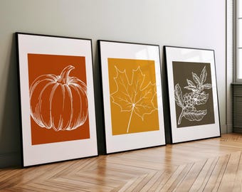 May include: Three framed illustrations of fall foliage. The first frame features a pumpkin in orange with a white outline on a rust-colored background. The second frame features a maple leaf in white on a yellow background. The third frame features a branch of berries in white on a dark brown background.
