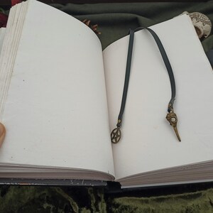 Puede incluir: Un libro abierto con p&aacute;ginas blancas en blanco, con un marcap&aacute;ginas de cuero oscuro con un dije de calavera de p&aacute;jaro de bronce y un dije de pentagrama. El libro est&aacute; sobre una superficie verde oscuro.