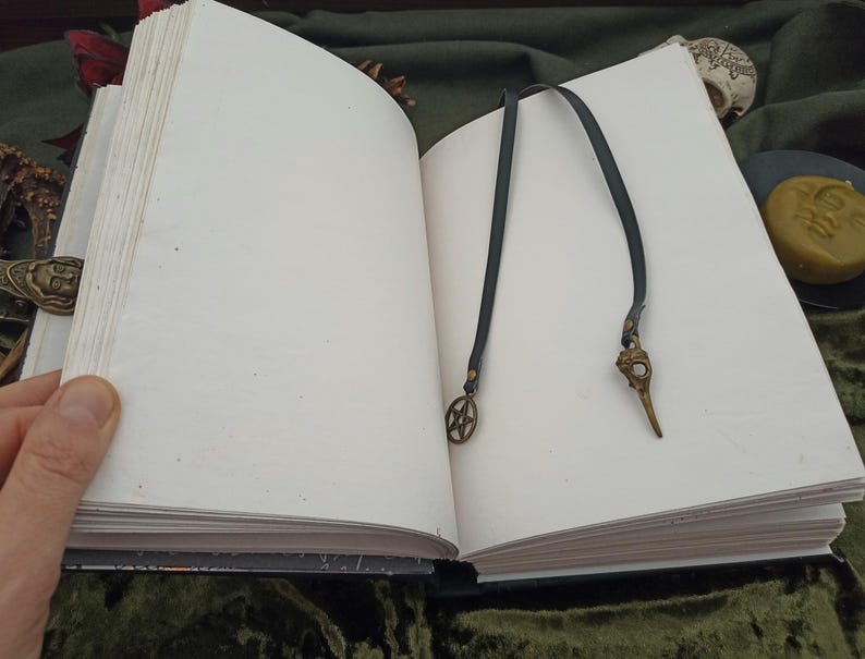 Puede incluir: Un libro abierto con p&aacute;ginas blancas en blanco, con un marcap&aacute;ginas de cuero oscuro con un dije de calavera de p&aacute;jaro de bronce y un dije de pentagrama. El libro est&aacute; sobre una superficie verde oscuro.