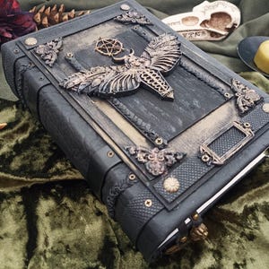 Puede incluir: Un libro encuadernado en cuero gris oscuro con adornos met&aacute;licos ornamentados. La portada presenta un gran dise&ntilde;o de polilla y pentagrama, con detalles decorativos en las esquinas. El libro est&aacute; cerrado, con un cierre visible en el borde.