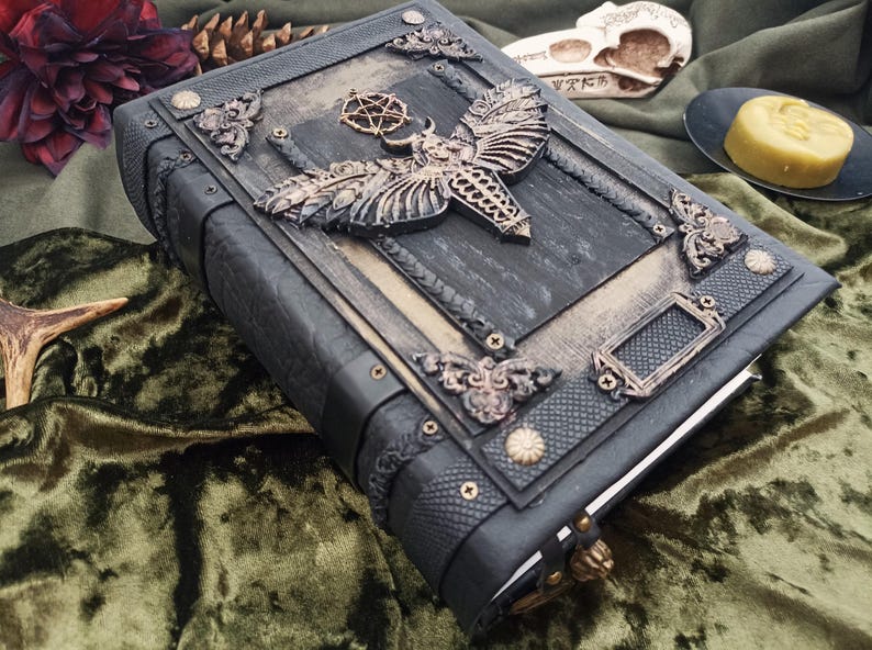 Puede incluir: Un libro encuadernado en cuero gris oscuro con adornos met&aacute;licos ornamentados. La portada presenta un gran dise&ntilde;o de polilla y pentagrama, con detalles decorativos en las esquinas. El libro est&aacute; cerrado, con un cierre visible en el borde.