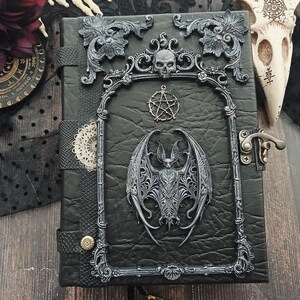 May include: Black leather-bound journal with ornate silver-colored embellishments. Features a bat, skull, and pentagram design. Includes a metal clasp and decorative frame. Gothic-themed.