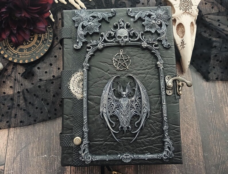 May include: Black leather-bound journal with ornate silver-colored embellishments. Features a bat, skull, and pentagram design. Includes a metal clasp and decorative frame. Gothic-themed.