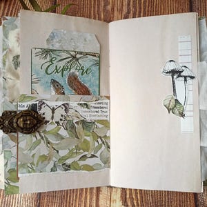 May include: Open journal with decorative elements. Features include a card with the word "Explore" and a squirrel illustration, a butterfly, and mushroom stickers. The journal is secured with a decorative clasp.