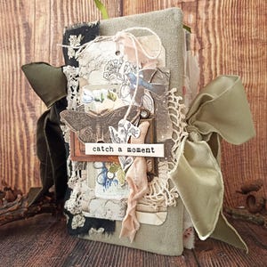May include: A handmade journal with a soft green cover, adorned with lace, ribbons, and decorative tags. The journal features a butterfly and floral design, with the words "catch a moment" on a small label. A large olive green ribbon is tied around the journal.