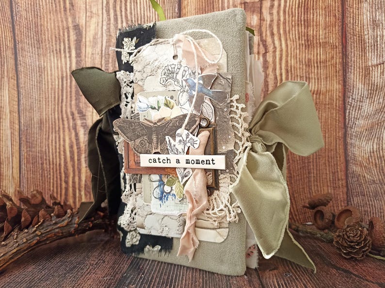 May include: A handmade journal with a soft green cover, adorned with lace, ribbons, and decorative tags. The journal features a butterfly and floral design, with the words "catch a moment" on a small label. A large olive green ribbon is tied around the journal.