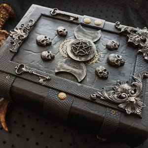 Peut inclure: Un livre de style gothique sombre avec des d&eacute;tails m&eacute;talliques orn&eacute;s. La couverture est orn&eacute;e de petits cr&acirc;nes, de croissants de lune et d'un pentagramme. Des cl&eacute;s et des coins d&eacute;coratifs en m&eacute;tal ajoutent &agrave; l'esth&eacute;tique vintage.