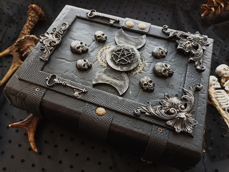 Peut inclure: Un livre de style gothique sombre avec des d&eacute;tails m&eacute;talliques orn&eacute;s. La couverture est orn&eacute;e de petits cr&acirc;nes, de croissants de lune et d'un pentagramme. Des cl&eacute;s et des coins d&eacute;coratifs en m&eacute;tal ajoutent &agrave; l'esth&eacute;tique vintage.