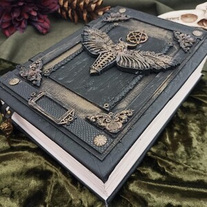 Puede incluir: Un libro oscuro y ornamentado con un dise&ntilde;o de polilla y pentagrama en relieve en la portada. El libro tiene una combinaci&oacute;n de colores negro y dorado con detalles met&aacute;licos decorativos. Las p&aacute;ginas son blancas y el libro parece estar encuadernado en cuero.
