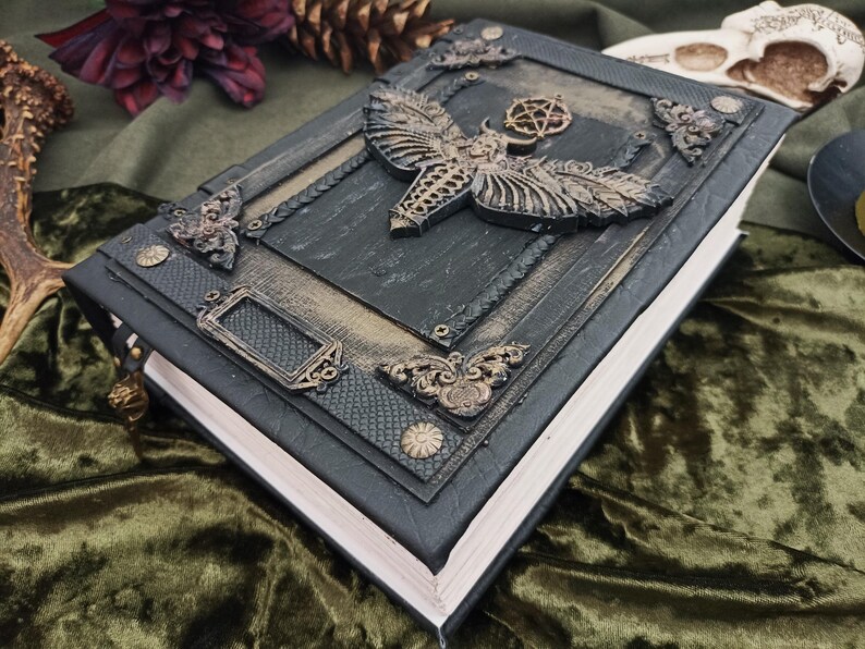 Puede incluir: Un libro oscuro y ornamentado con un dise&ntilde;o de polilla y pentagrama en relieve en la portada. El libro tiene una combinaci&oacute;n de colores negro y dorado con detalles met&aacute;licos decorativos. Las p&aacute;ginas son blancas y el libro parece estar encuadernado en cuero.