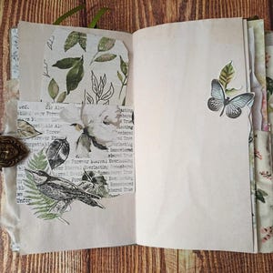 May include: Open journal with blank pages and decorative elements. Features a butterfly sticker, floral and leaf prints, and a bird illustration. Includes a decorative clasp and a green ribbon bookmark. The pages have a vintage, aged appearance.