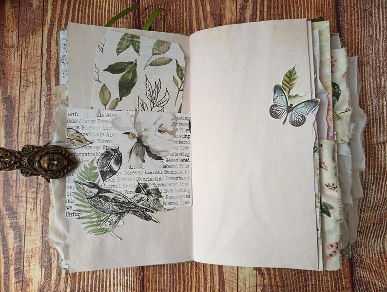 May include: Open journal with blank pages and decorative elements. Features a butterfly sticker, floral and leaf prints, and a bird illustration. Includes a decorative clasp and a green ribbon bookmark. The pages have a vintage, aged appearance.