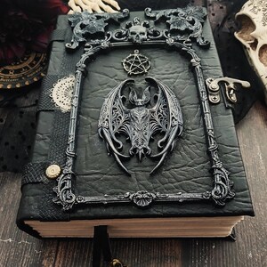 May include: A black leather-bound journal with a decorative frame featuring a bat, skull, and pentagram. The journal has a metal clasp and a black ribbon bookmark. The cover has a textured appearance.