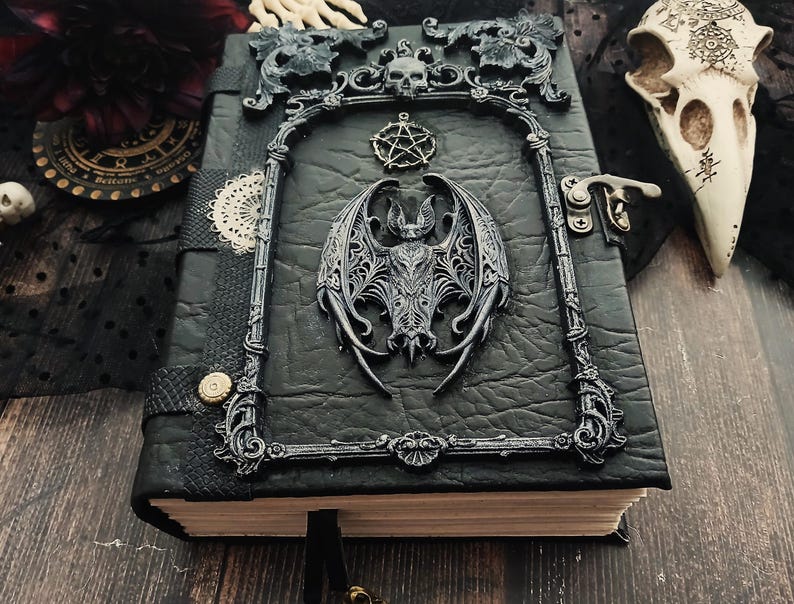 May include: A black leather-bound journal with a decorative frame featuring a bat, skull, and pentagram. The journal has a metal clasp and a black ribbon bookmark. The cover has a textured appearance.