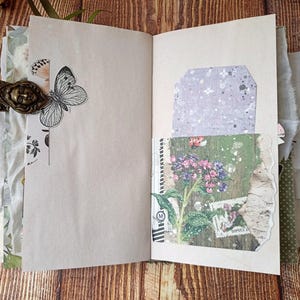 May include: Open journal with blank pages, adorned with a butterfly illustration and a decorative metal clasp. Features a tag and a pocket with floral and botanical imagery. The journal is on a wooden surface.