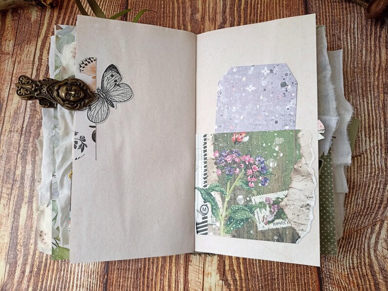 May include: Open journal with blank pages, adorned with a butterfly illustration and a decorative metal clasp. Features a tag and a pocket with floral and botanical imagery. The journal is on a wooden surface.