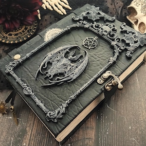 May include: A black leather-bound journal with ornate, dark gray metal embellishments. The cover features a bat, a pentagram, and a decorative frame with skulls. A metal clasp secures the journal, and a sun charm hangs from a black ribbon.