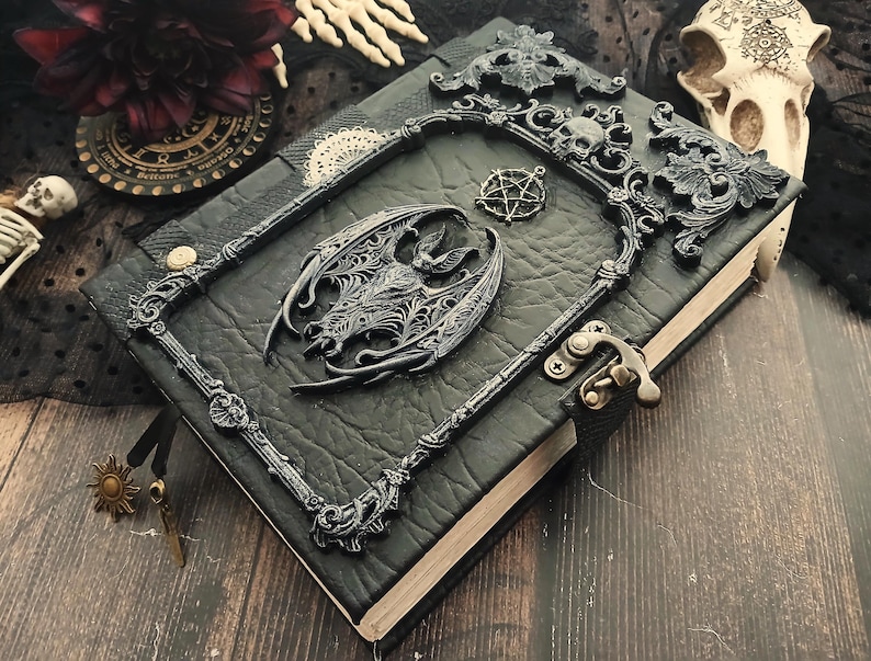 May include: A black leather-bound journal with ornate, dark gray metal embellishments. The cover features a bat, a pentagram, and a decorative frame with skulls. A metal clasp secures the journal, and a sun charm hangs from a black ribbon.