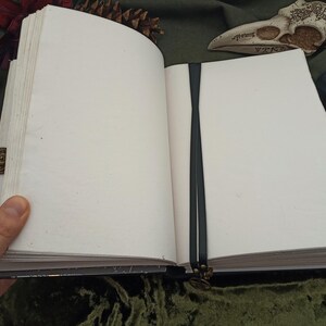 Puede incluir: Un diario abierto con una cinta de lectura negra. Las p&aacute;ginas son gruesas y blanquecinas. La cubierta es oscura con un cierre decorativo. El diario est&aacute; abierto, revelando p&aacute;ginas en blanco.