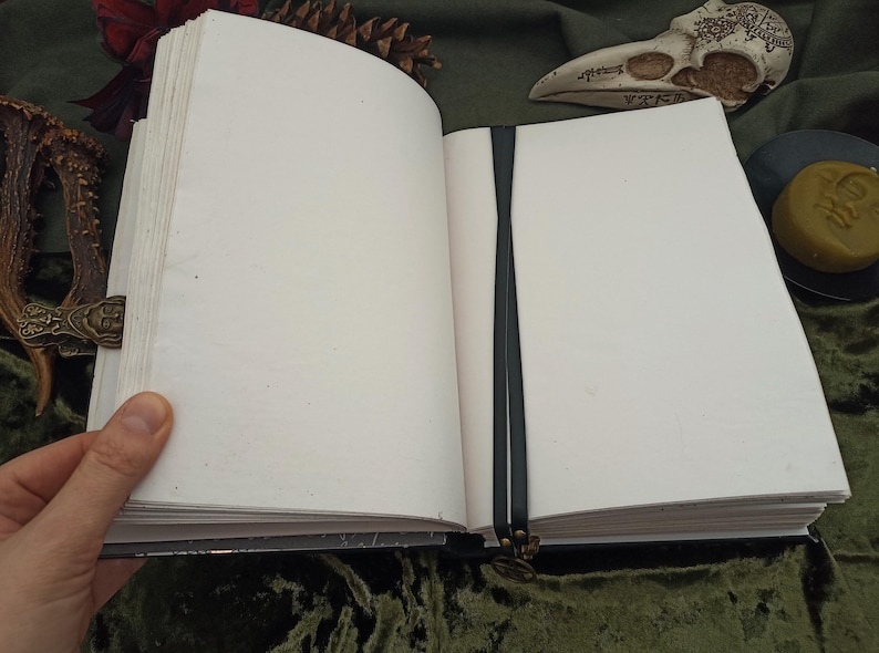 Puede incluir: Un diario abierto con una cinta de lectura negra. Las p&aacute;ginas son gruesas y blanquecinas. La cubierta es oscura con un cierre decorativo. El diario est&aacute; abierto, revelando p&aacute;ginas en blanco.