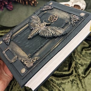 Puede incluir: Un libro verde oscuro en relieve con un dise&ntilde;o de polilla y pentagrama. El libro tiene adornos met&aacute;licos dorados en la portada y el lomo. Las p&aacute;ginas son blancas y el libro est&aacute; sostenido en una mano.