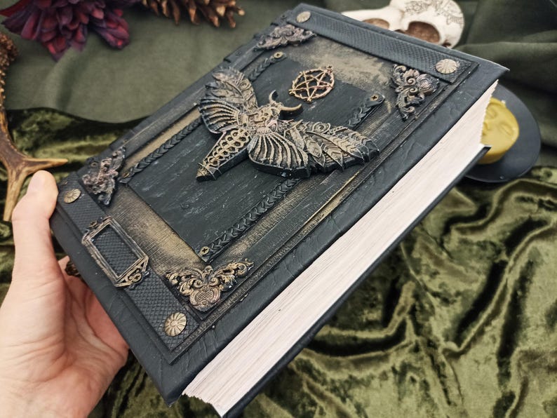 Puede incluir: Un libro verde oscuro en relieve con un dise&ntilde;o de polilla y pentagrama. El libro tiene adornos met&aacute;licos dorados en la portada y el lomo. Las p&aacute;ginas son blancas y el libro est&aacute; sostenido en una mano.